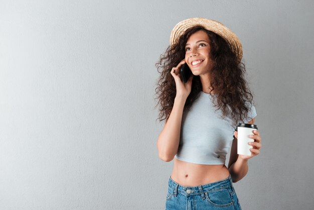 Lächelnde lockige Frau im Hut, die Kaffee trinkt und durch das Smartphone spricht