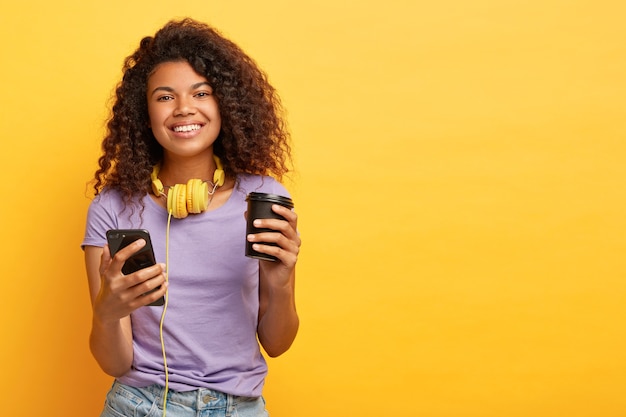 Lächelnde lockige Frau sieht Video auf Handy während der Kaffeepause, hört Audiospuren über Kopfhörer, hat gute Laune, trägt lässiges Outfit, isoliert über gelbem Hintergrund, Leerzeichen