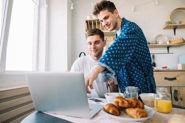 Lächelnde Männer, die auf Laptop auf Tabelle mit Hörnchenbrot und Glas Saft zeigen