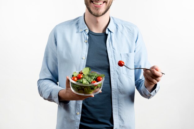 Lächelnde Mannholdinggabel mit Kirschtomate und Schüssel Salat gegen weißen Hintergrund