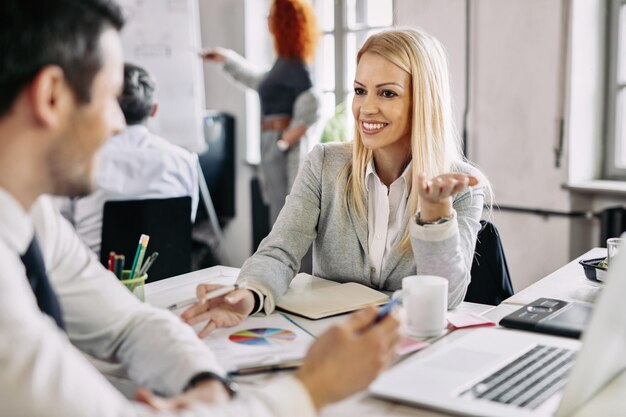 Lächelnde mittlere erwachsene Geschäftsfrau, die mit ihrem männlichen Kollegen im Büro spricht und eine neue Geschäftsstrategie entwickelt. Es gibt Menschen im Hintergrund