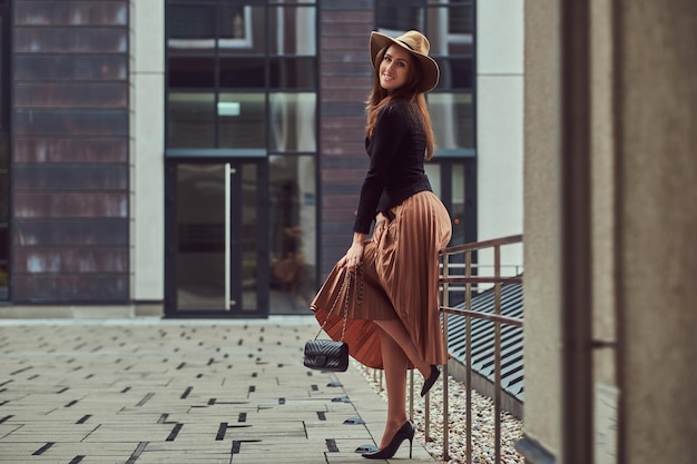 Lächelnde, modische, elegante Frau, die eine schwarze Jacke, einen braunen Hut und einen Rock mit einer Handtaschenkupplung trägt und posiert, während sie sich auf ein Stahlgeländer in einem europäischen Stadtzentrum lehnt.