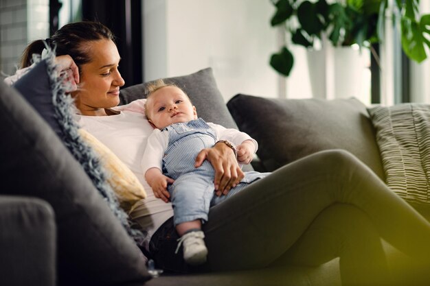 Lächelnde Mutter, die ihren kleinen Jungen ansieht, während sie ihn hält und sich im Wohnzimmer entspannt