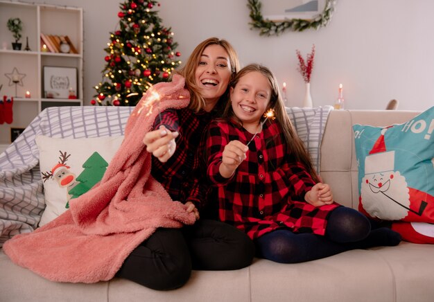 Lächelnde Mutter und Tochter halten Wunderkerzen bedeckt mit Decke, die auf Couch sitzt und Weihnachtszeit zu Hause genießt