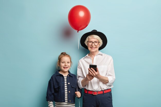 Lächelnde Oma in schwarzem stilvollem Hut, weißem elegantem Hemd und formeller Hose, hält Handy, weiß, wie man moderne Geräte gut benutzt, feiert Geburtstag des kleinen Kindes, das roten Luftballon hält