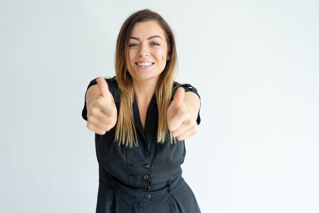 Lächelnde recht junge Frau im schwarzen Kleid, das Thumbs-up beim Ausdrücken ihrer Zustimmung zeigt