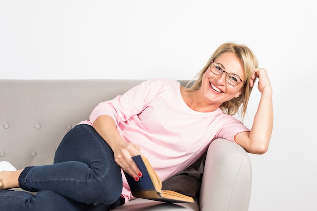 Lächelnde reife Frau, die sich auf dem Sofa hält Buch in der Hand am weißen Hintergrund lehnt