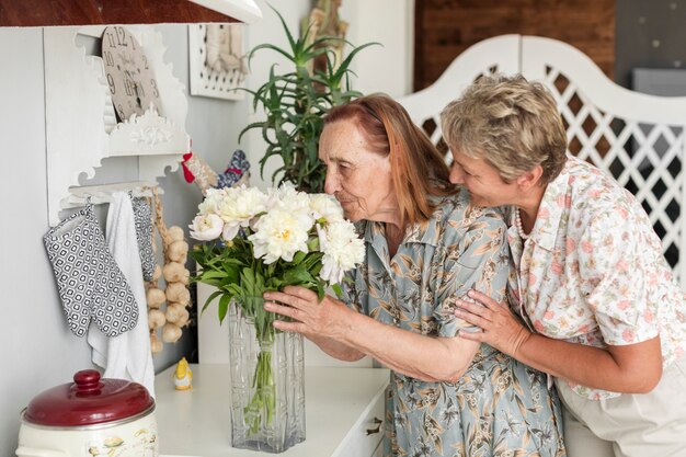 Lächelnde reife Frau, die zu Hause ihren riechenden Vase der weißen Blumen der Mutter betrachtet