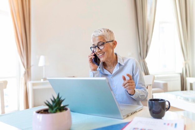 Lächelnde, reife, schöne Geschäftsfrau mit weißem Haar, die im hellen, modernen Heimbüro am Laptop arbeitet