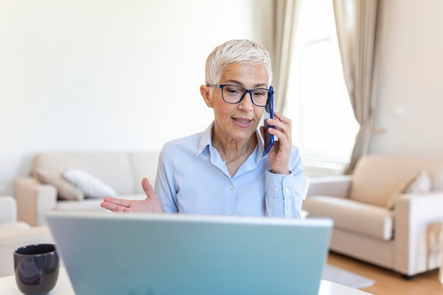 Lächelnde, reife, schöne Geschäftsfrau mit weißen Haaren, die im hellen, modernen Heimbüro am Laptop arbeitet