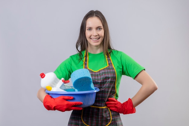 Lächelnde Reinigung junges Mädchen, das Uniform in roten Handschuhen hält, die Reinigungswerkzeuge halten, legte ihre Hand auf Hüfte auf lokalisiertem weißem Hintergrund