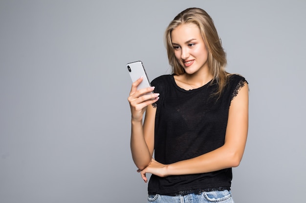 Lächelnde reizende junge Frau stehend und mit Handy über grauem Hintergrund