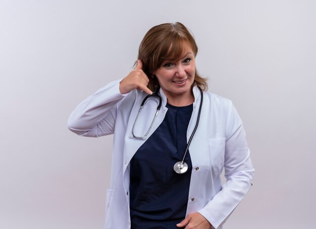 Lächelnde Ärztin mittleren Alters, die medizinische Robe und Stethoskop trägt, die Telefongeste auf isolierter weißer Wand tut