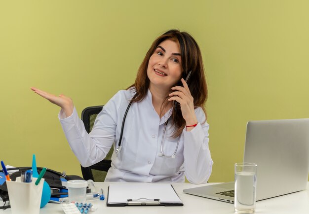 Lächelnde Ärztin mittleren Alters, die medizinische Robe und Stethoskop trägt, sitzt am Schreibtisch mit medizinischen Werkzeugen und Laptop, die am Telefon sprechen, das leere Hand lokalisiert zeigt