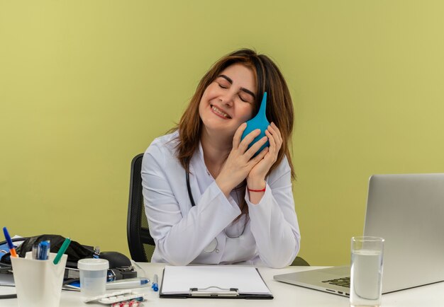 Lächelnde Ärztin mittleren Alters, die medizinische Robe und Stethoskop trägt, sitzt am Schreibtisch mit medizinischen Werkzeugen und Laptop, die Einlauf nahe Kopf mit geschlossenen Augen lokalisiert halten