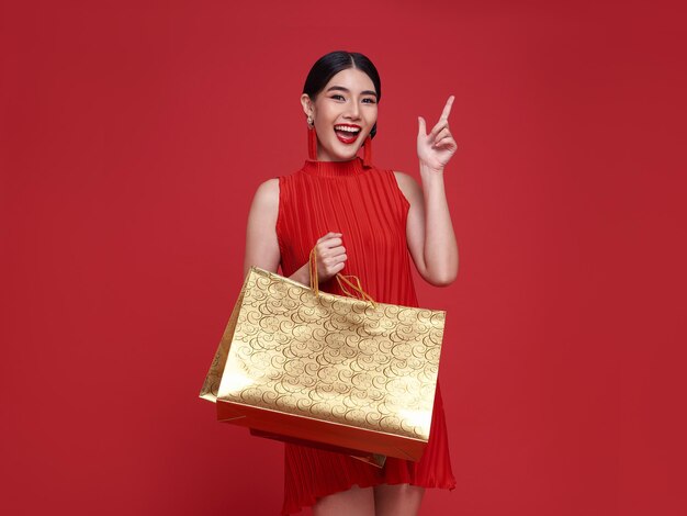 Lächelnde schöne asiatische Frau, die Einkaufstaschen hält und auf den Kopienraum zeigt, der auf rotem Hintergrund für das Verkaufskonzept des chinesischen Neujahrs isoliert ist.
