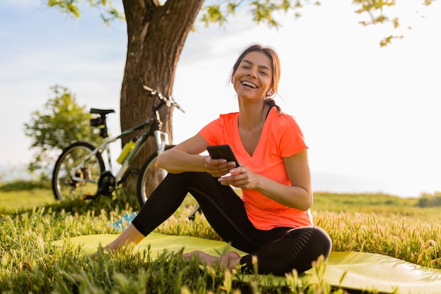 Lächelnde schöne frau, die telefon hält, das sport am morgen im park tut