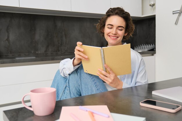 Lächelnde schöne Frau sitzt mit einem Notizbuch in der Küche, liest Notizen, studiert, macht Hausaufgaben