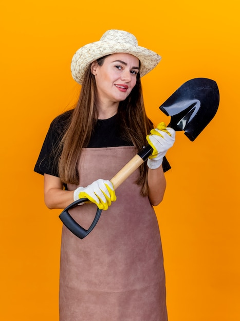 Lächelnde schöne Gärtnerin, die Uniform und Gartenhut mit Handschuhen hält, die Spaten lokalisiert auf Orange halten