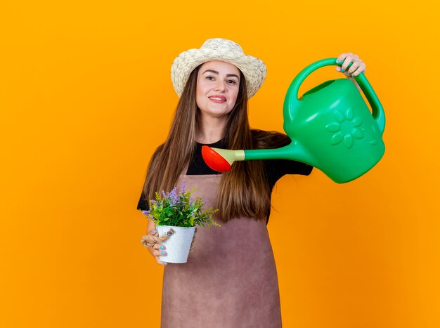 Lächelnde schöne Gärtnerin, die Uniform und Gartenhut trägt, die Blume im Blumentopf mit Bewässerungsdose tränkt, lokalisiert auf orange Hintergrund