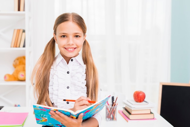 Lächelnde Schulmädchenschreibensaufgabe mit Bleistift im Notizbuch am Klassenzimmer