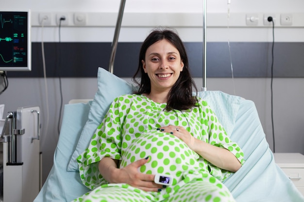 Kostenloses Foto lächelnde schwangere frau liegt im bett und hält die hände auf dem bauch in der krankenstation. fröhliche patientin mit bauchschwangerschaft blickt in die kamera und wartet auf den arzt, der während des kontrollbesuchs eine untersuchung durchführt