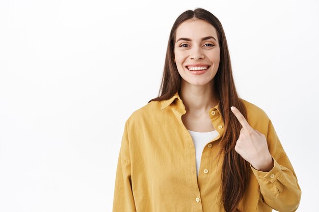 Lächelnde selbstbewusste Frau, die mit stolzem, entschlossenem Gesicht auf sich selbst zeigt, sich selbst fördert, ihre neuen weißen Zähne zeigt und über der Studiowand steht