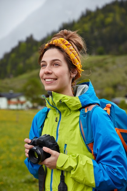 Lächelnde sorglose europäische Frau hält professionelle Kamera, schaut positiv in die Ferne