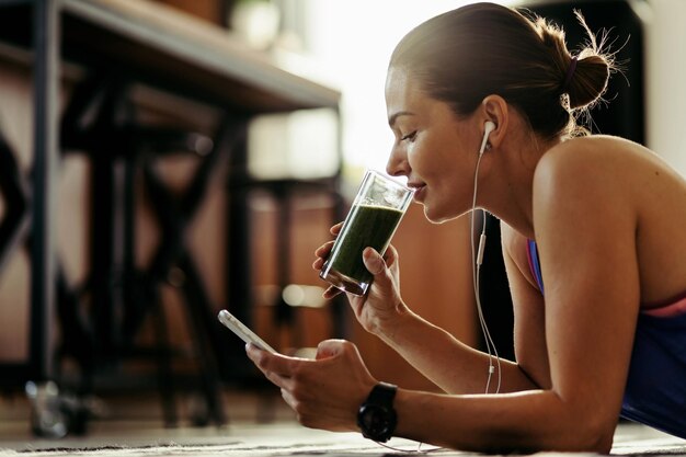 Lächelnde Sportlerin benutzt Smartphone und trinkt gesunden Smoothie, während sie sich zu Hause auf den Boden legt