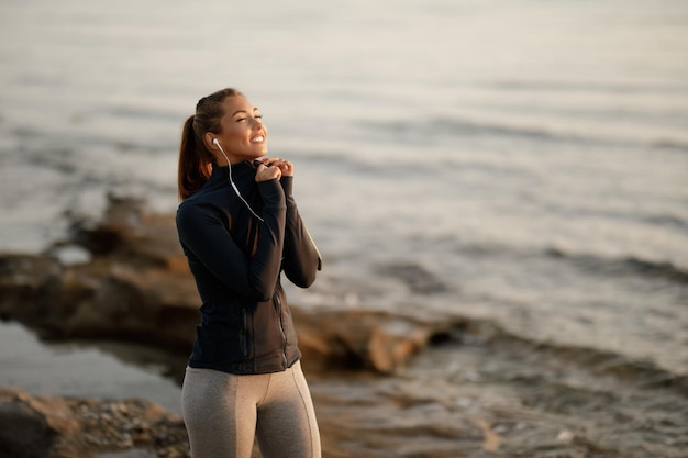 Lächelnde sportliche Frau, die mit geschlossenen Augen an der Küste steht, während sie Musik über Kopfhörer genießt Kopieren Sie Platz
