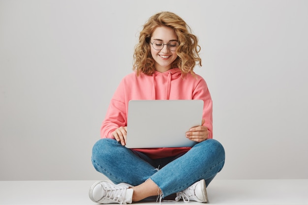 Lächelnde Studentin, die Aufsatz schreibt und mit Laptop auf Boden sitzt