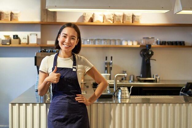 Lächelnde Studentin, die Teilzeit im Café Barista arbeitet, zeigt Daumen hoch, trägt Schürze, steht in der Nähe von Kaffee