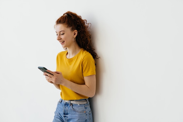 Lächelnde Studentin mit roten Haaren, die ein Handy benutzt, das glücklich in die Kamera schaut, die über weißem Hintergrund steht