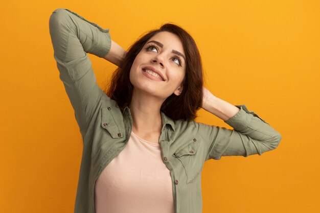 Lächelnde Suche nach jungem schönem Mädchen, das olivgrünes T-Shirt hält Hände auf hinter Kopf lokalisiert auf gelber Wand