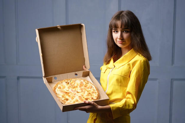 Lächelnde süße junge Frau in einem gelben Overall, der Pizza liefert.