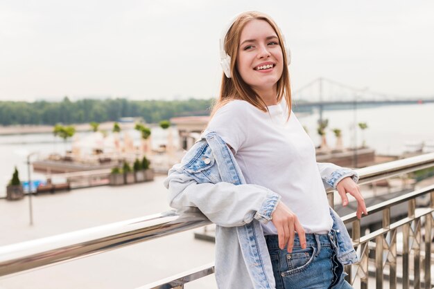 Lächelnde tragende hörende Musik des Kopfhörers der Frau, die nahe metallischem Geländer steht