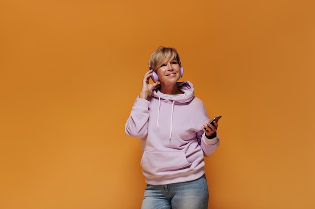 Lächelnde trendige alte Dame mit blonder kühler Frisur im rosa Sweatshirt und in den hellen Jeans, die mit lila Kopfhörern und Smartphones aufwerfen.