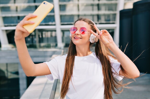 Lächelnde und tanzende junge Frau, die mit ihrem Smartphone ein Selfie macht und Musik in Kopfhörern hört