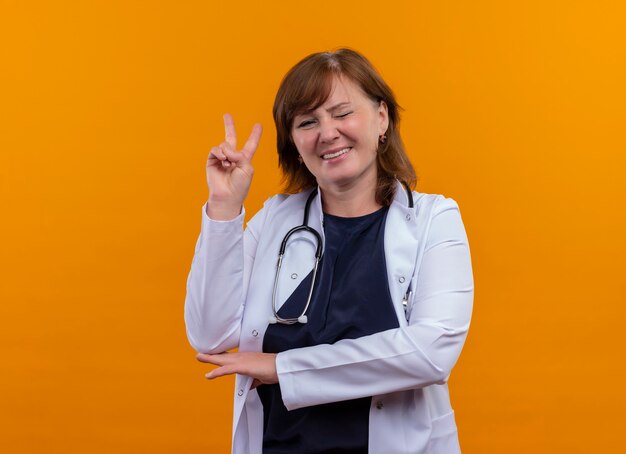 Lächelnde und zwinkernde Ärztin mittleren Alters, die medizinische Robe und Stethoskop trägt, die Friedensgeste auf isolierter orange Wand mit Kopienraum tun