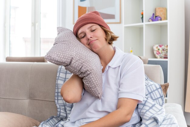 Lächelnde ungesunde junge Frau in warmem Hut mit Decke, die an Erkältung und Grippe leidet, Kissen hält Kopf auf Kissen mit geschlossenen Augen auf der Couch im hellen Wohnzimmer sitzend