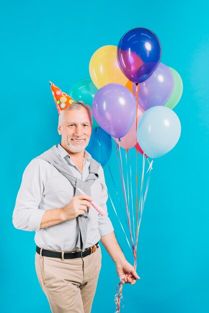 Lächelnder älterer Mann mit den Ballonen und Partyhorn, die Kamera auf blauem Hintergrund betrachten