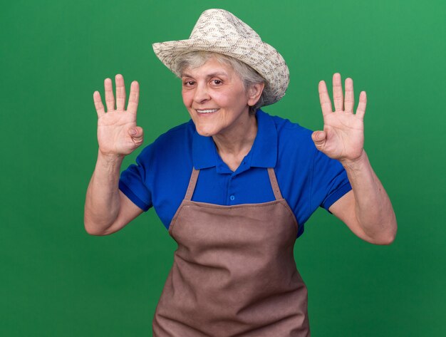 Lächelnder älterer weiblicher Gärtner, der Gartenhut trägt, der vier mit den Fingern lokalisiert auf grüner Wand mit Kopienraum gestikuliert