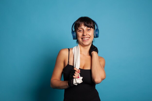 Lächelnder aktiver trainer mit kopfhörern, der musik hört, während er während des pilates-trainings körpermuskeln trainiert. athletische frau, die im studio mit blauem hintergrund an körperausdauer arbeitet