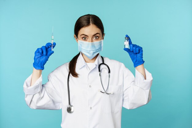 Lächelnder Arzt des medizinischen Personals mit Gesichtsmaske und Gummihandschuhen, der Spritze und Impfstoff von covid p zeigt