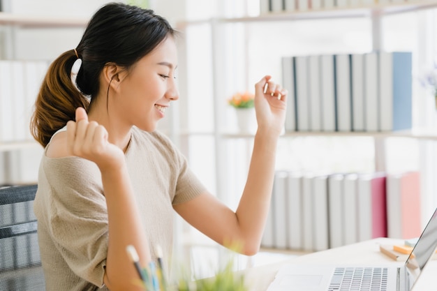 Lächelnder asiatischer Frauenarbeitslaptop der schönen Junge auf Schreibtisch im Wohnzimmer zu Hause