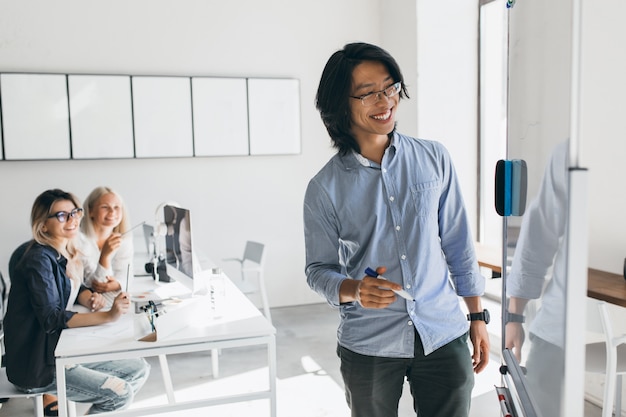 Kostenloses Foto lächelnder asiatischer freiberuflicher entwickler, der aktionsplan des flipcharts zeichnet. blonde junge managerinnen, die ausländische kollegin betrachten, die etwas an bord schreibt.