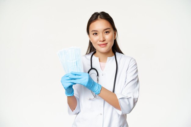 Lächelnder asiatischer Hausarzt, der sterile Gesichtsmasken zeigt, die verhindern, dass Covid stehend ...