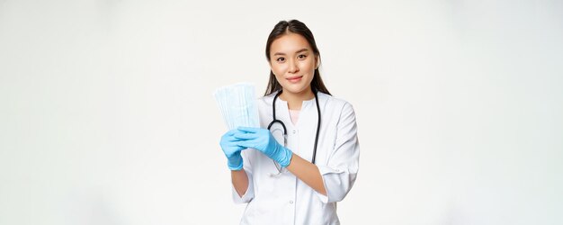 Lächelnder asiatischer Hausarzt, der sterile Gesichtsmasken zeigt, die verhindern, dass covid19 in Uniform vor weißem Hintergrund steht