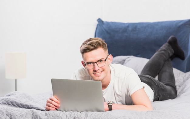 Lächelnder attraktiver junger Mann, der auf Bett unter Verwendung der digitalen Tablette liegt