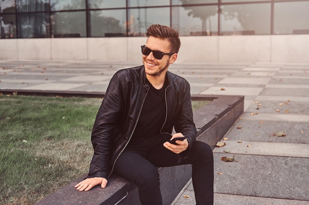 Lächelnder attraktiver Mann chattet auf dem Handy, während er draußen in der Nähe des Gebäudes sitzt.
